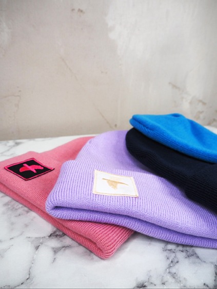 italienische mode kleiber werksverkauf Stapel bunter Beanies: Pink, Lavendel, Marineblau und Blau. Jede Beanie hat einen Logo-Aufnäher.