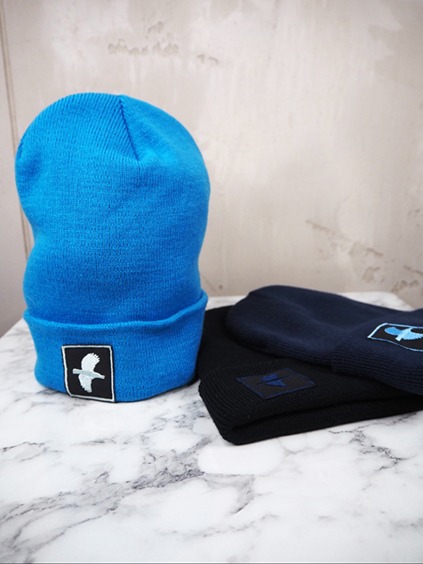 italienische mode kleiber werksverkauf Blaue Beanie mit Vogellogo, neben marineblauen und schwarzen Beanies auf einer Marmoroberfläche.