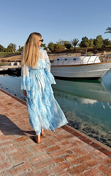 italienische mode kleiber werksverkauf Frau in blauem Kleid spaziert am Wasser entlang, Boot im Hintergrund